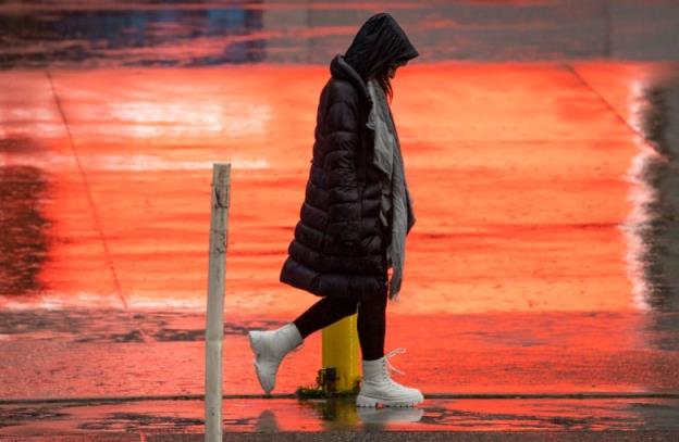 A woman in a long jacket walks down a wet street with her head down.  