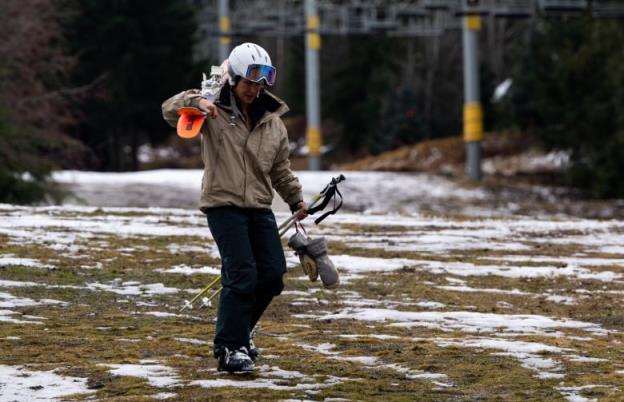 A skier holding skis, poles and gloves walks down a slope of grass and patchy snow.