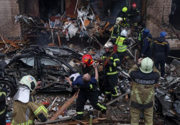 A firefighter carries a disabled man out of his apartment block after it was hit in a Russian air raid. He is walking around piles of debris and a destroyed car. Other firefighters are behind him .