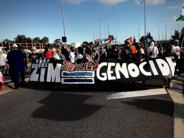 Protesters walking with a giant banner saying ZIM=Genocide. They are carrying the banner in front of them as they walk forward