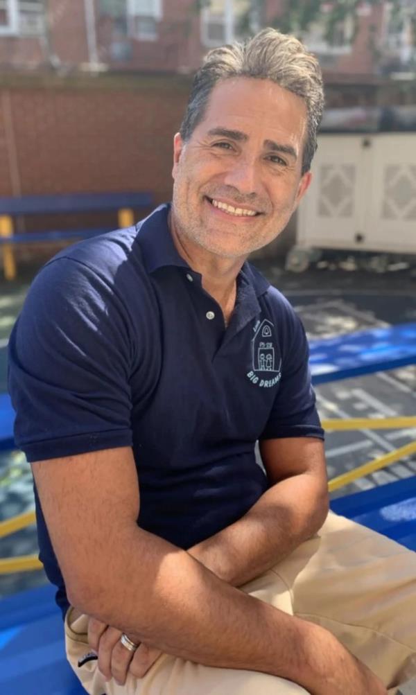 Robert Quintana, principal of PS 28 in Queens, sitting down, smiling into the camera, wearing a dark blue t-shirt and brown pants