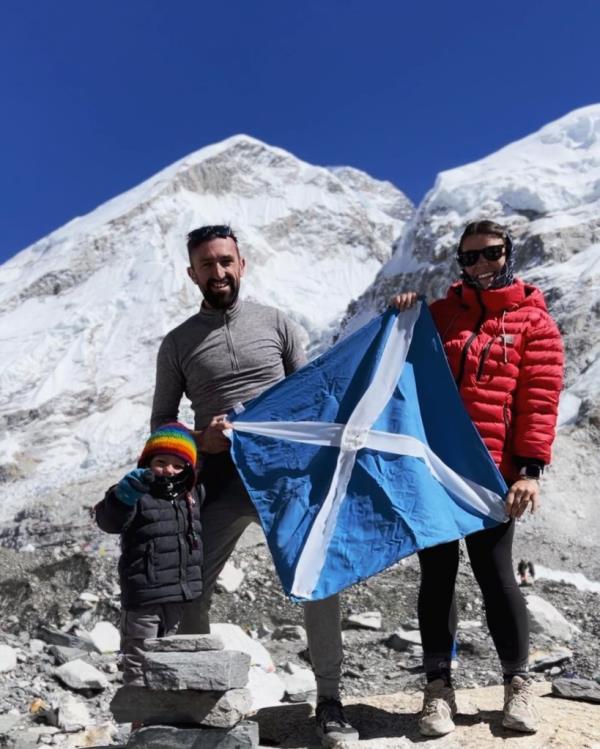 The Dallas family and others hold a flag in front of a mountain while trekking to Everest ba<em></em>se camp.