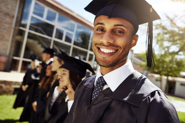 Smiling college gradudate wearing cap and gown with classmates in background.