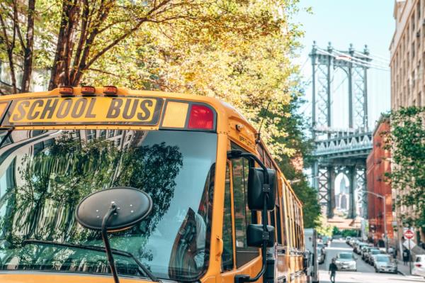 Yellow school bus driving in Brooklyn with the Manhattan bridge in the background.