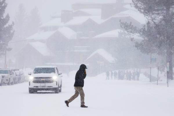 A person crosses a snow-covered road in Mammoth Lakes, California, with a white SUV traveling nearby and a large building with snow on the roof in the background.