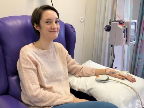 Emma Barker in a hospital chair, with tubes going into her left hand/arm, propped on a pillow.