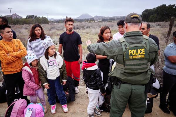 Migrants are processing near the U.S.-Mexico border, with police and people standing together in a field.