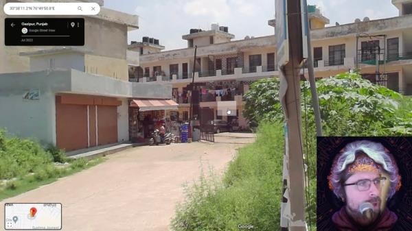 A still from a YouTube video showing Google streetview of a building in India with a street and greenery in the foreground and bottom right a man narrating the video 